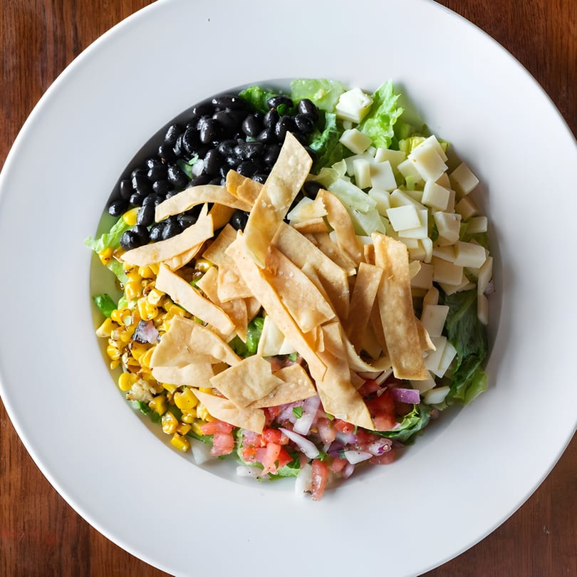 Overhead view of a colorful salad with black beans, corn, tortilla strips, cheese, and pico de gallo.