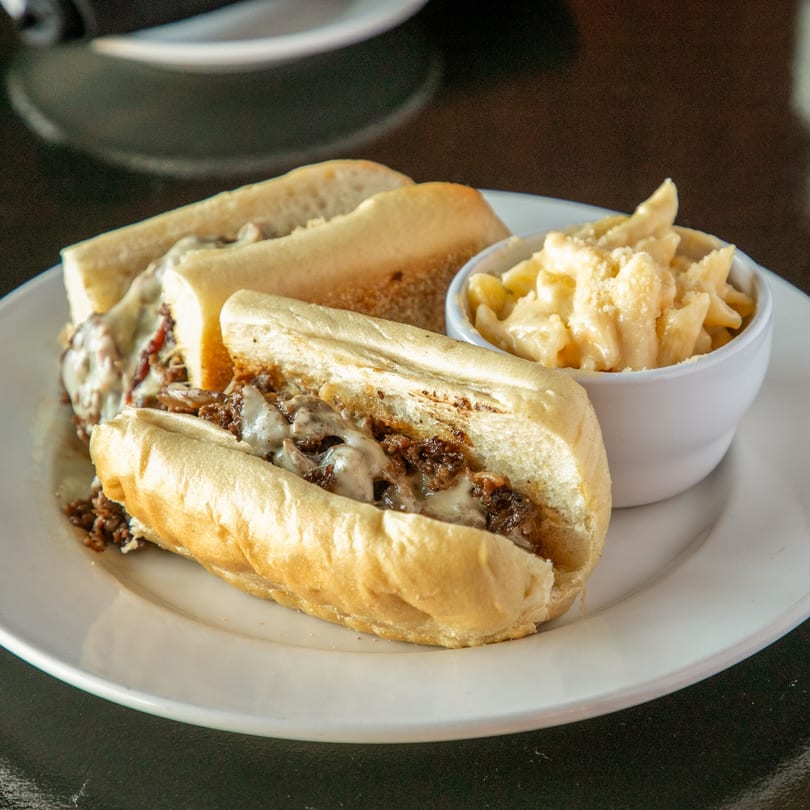 Two cheesesteak sandwiches with melted cheese and a side of macaroni and cheese.